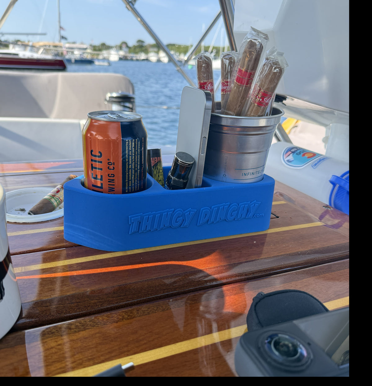 A blue silicone drink holder organizer on a wooden table of a boat, holding cigars in a metal cup, an orange can, and other small items.